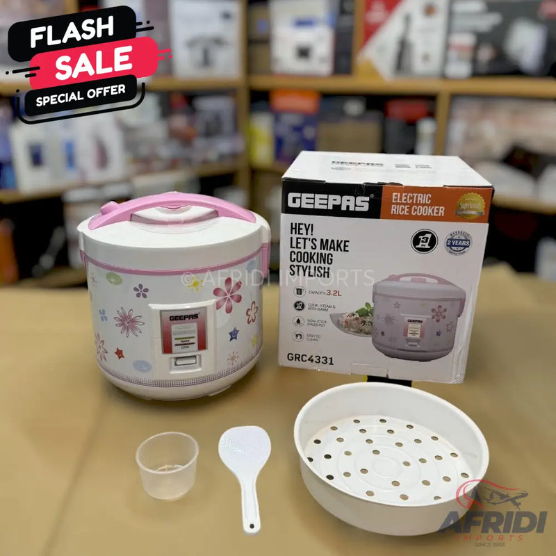 A white electric rice cooker with a pink lid and floral pattern, accompanied by its packaging, a measuring cup, a serving spoon, and a steaming basket.