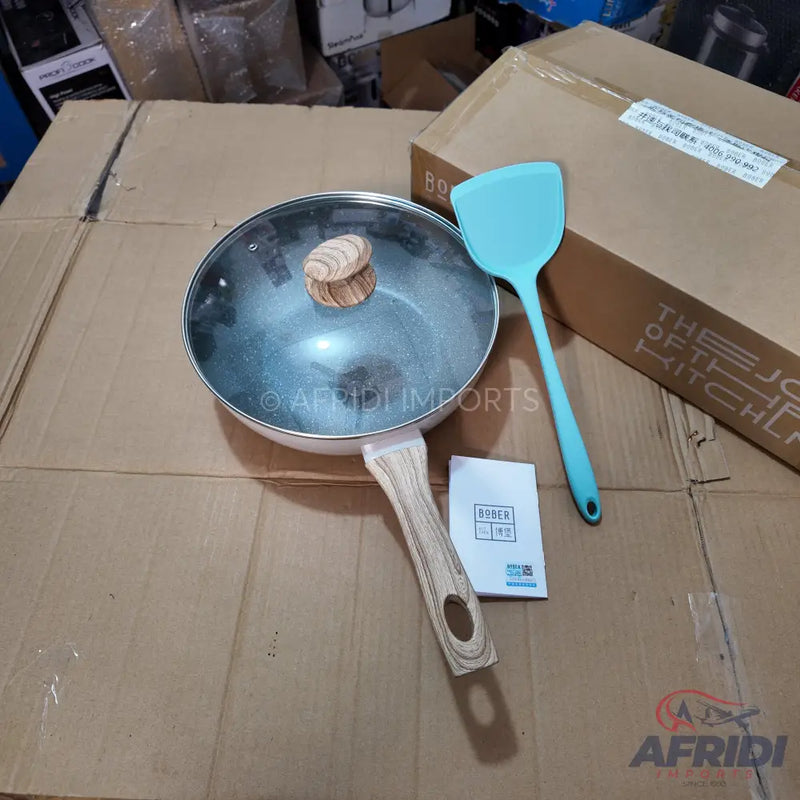 A white non-stick frying pan with a wooden handle and glass lid, accompanied by a blue silicone spatula and an instruction booklet.
