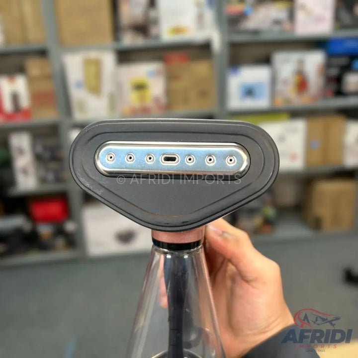 Handheld garment steamer with a dark grey head featuring a light blue-grey steam plate with multiple holes and a central USB-C port, attached to a clear plastic water reservoir.