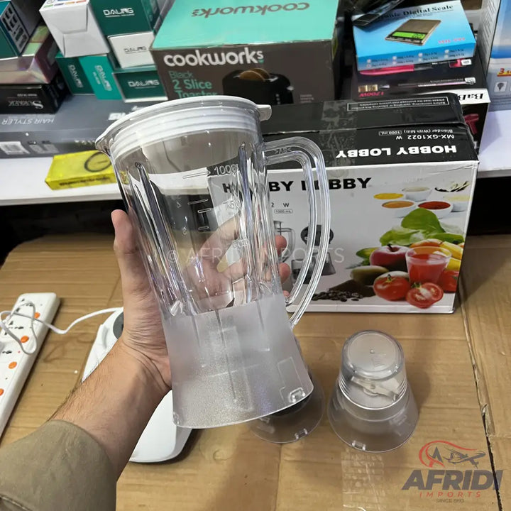 A clear plastic blender jar with a lid, held by a hand, alongside a smaller blending attachment.