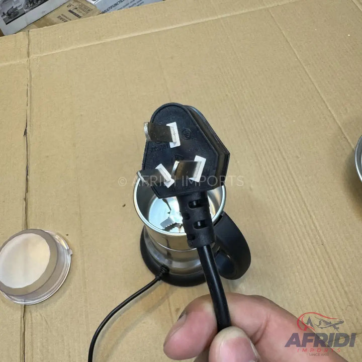 A black three-pin UK-style electrical plug attached to a power cord, held in a hand, with a small silver and black electric grinder in the background.