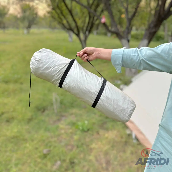 Rolled-up light-colored camping mat or tent in a carry bag with black straps.