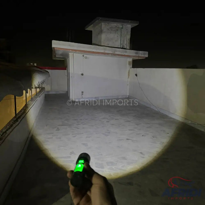 Handheld flashlight with a green illuminated switch, casting a bright beam on a concrete rooftop at night.