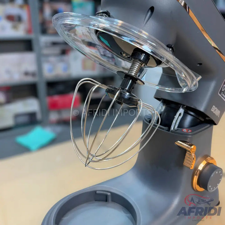 Dark gray stand mixer with a clear splash guard, a wire whisk attachment, and gold accents.