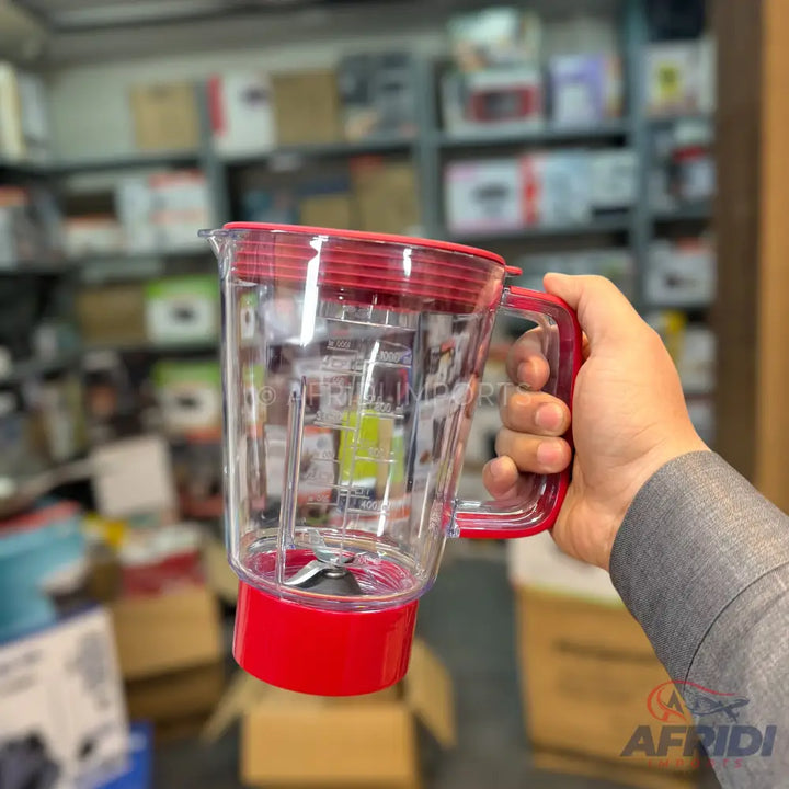 A transparent blender pitcher with a red lid, handle, and base, featuring internal measurement markings and a two-blade assembly at the bottom.