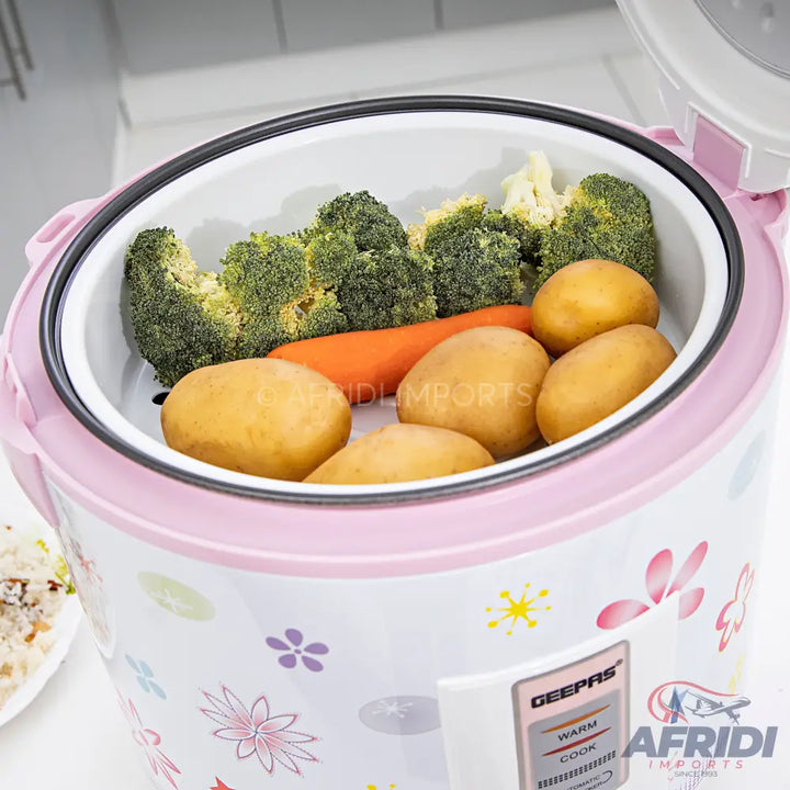 A pink and white electric rice cooker with a floral pattern, featuring a steaming basket filled with broccoli, carrots, and potatoes.