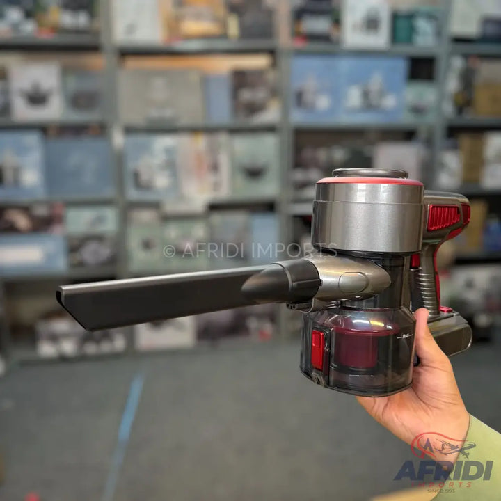 A cordless handheld vacuum cleaner with a grey and red body, a transparent dustbin, and an attached crevice tool.