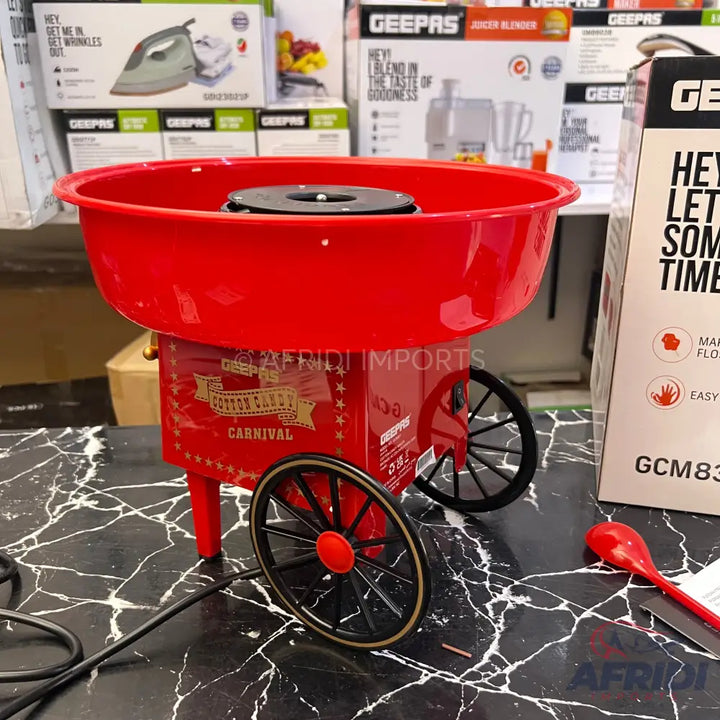 A red carnival-style cotton candy maker with black wheels and a red measuring spoon.