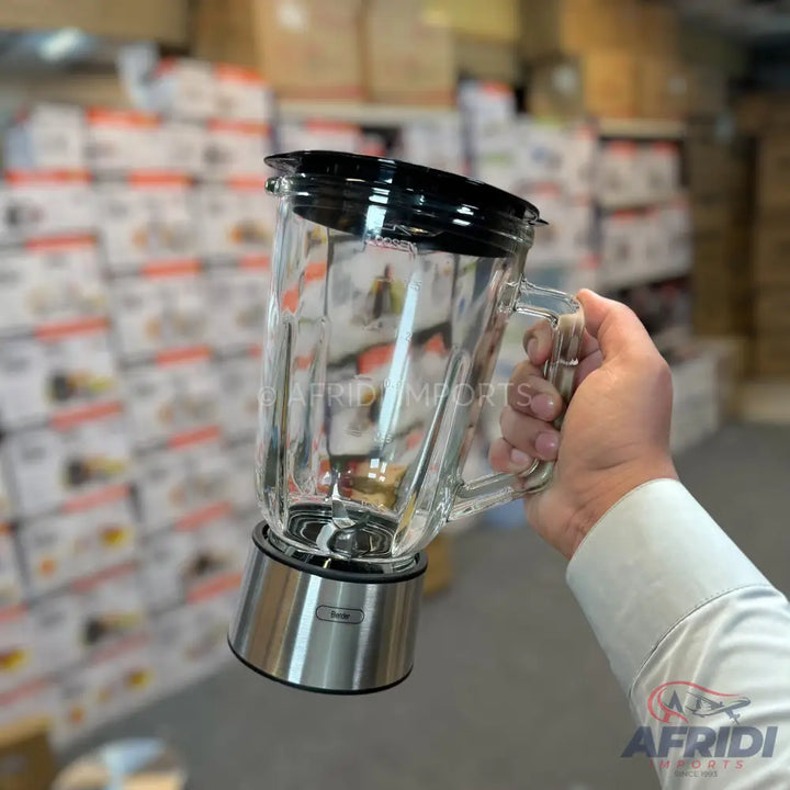A clear glass blender jar with a black lid and a shiny silver base, held by a hand.