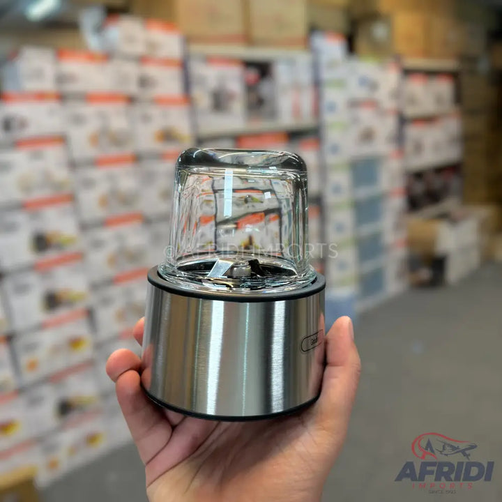 A handheld food chopper with a stainless steel base and a clear glass cup, featuring a visible blade.