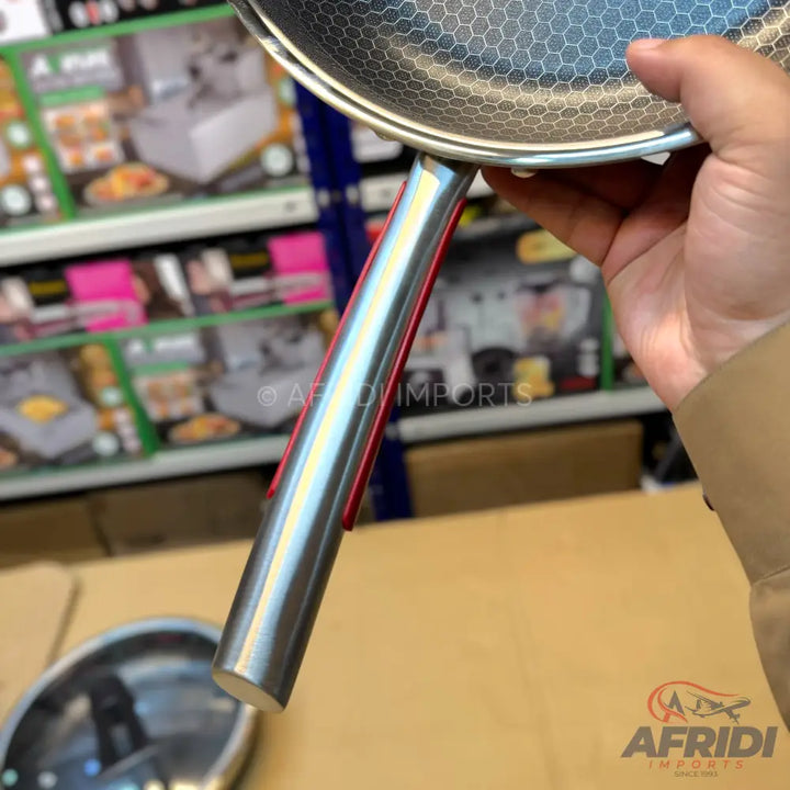 A hand holding a stainless steel frying pan with a honeycomb textured cooking surface and a red-accented handle.