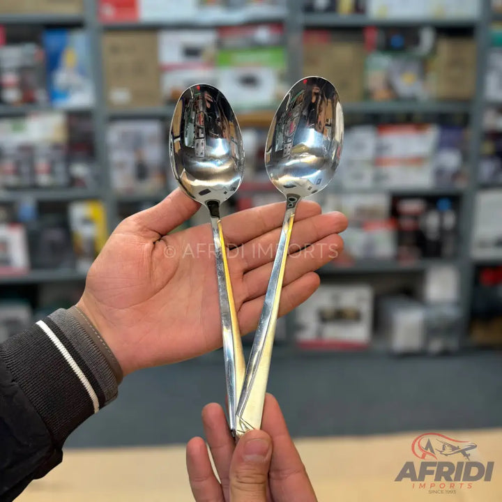 Two hands holding a pair of polished, silver-colored serving spoons with gold-colored accents on their handles.