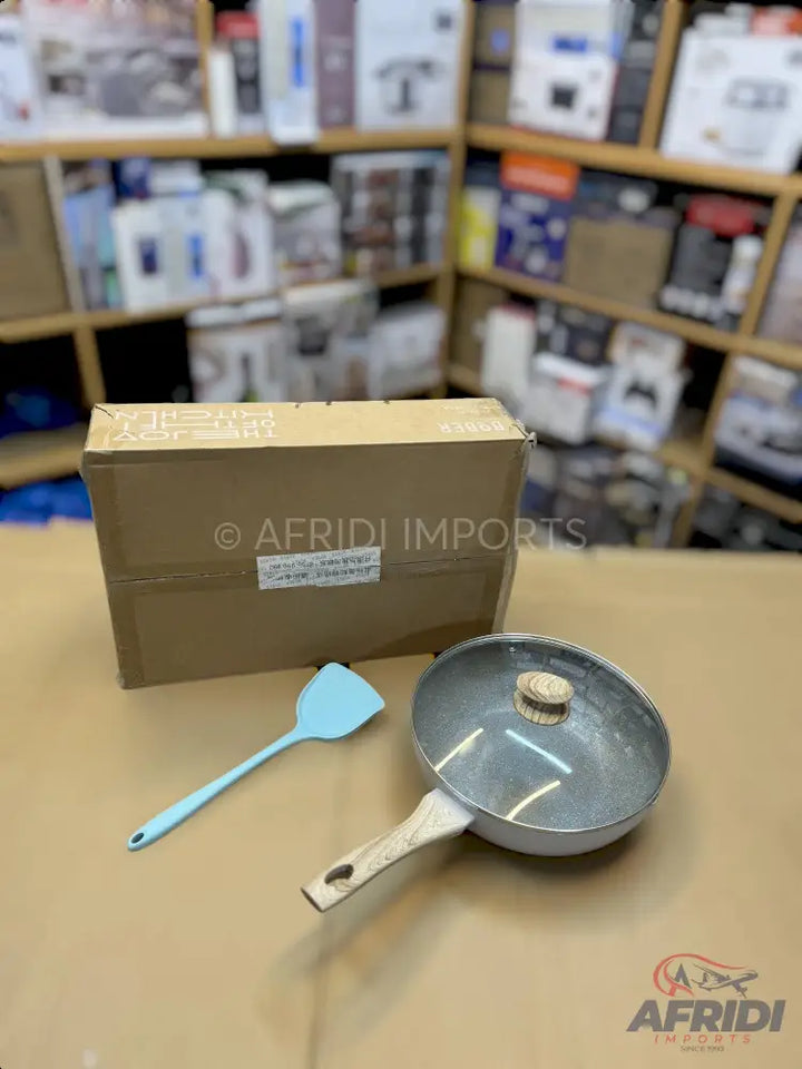 A silver frying pan with a glass lid and a wooden handle, accompanied by a light blue silicone spatula, and a brown cardboard box.