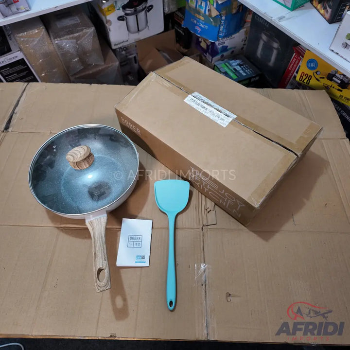 A silver wok with a wooden handle and a glass lid, accompanied by a blue silicone spatula and a small instruction manual.