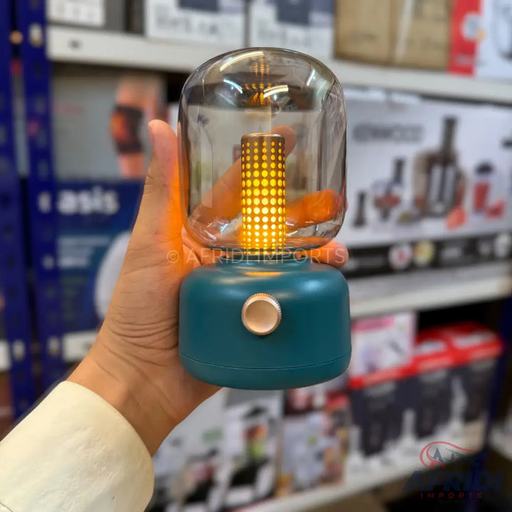 A portable teal and bronze lantern with a clear glass dome, illuminating with warm yellow LED lights.