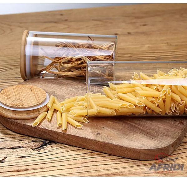 Two clear glass cylindrical food storage containers with wooden lids, one filled with dry pasta and the other with dried herbs.
