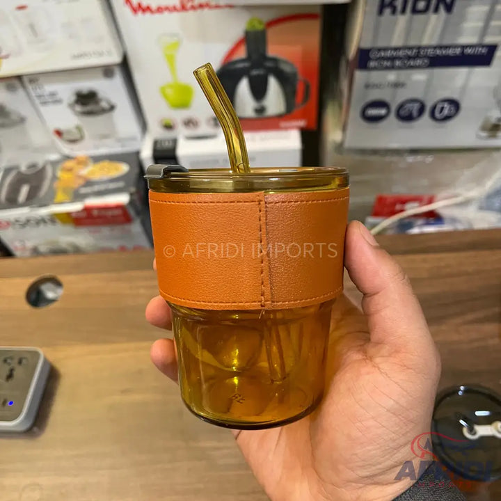 A clear amber-colored glass cup with a lid, a reusable straw, and an orange faux leather sleeve.