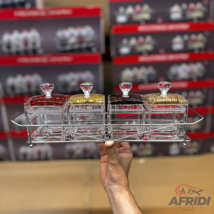 A set of four clear acrylic spice jars with crystal-like knob lids, held in a silver metal rack.