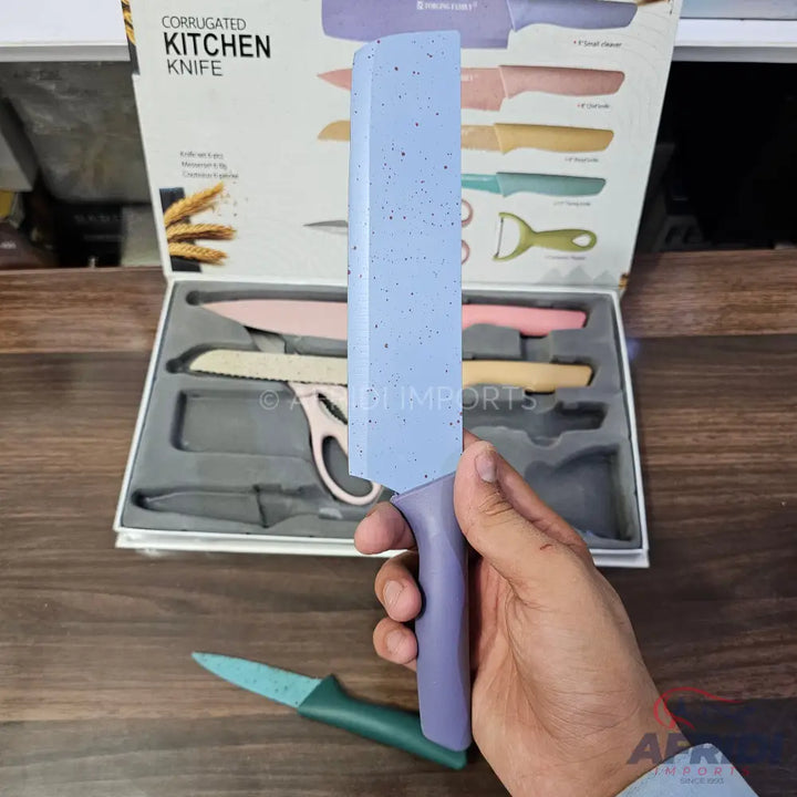 A set of colorful kitchen knives with speckled blades and matching handles, including a peeler and scissors, packaged in a display box, with one knife being held.