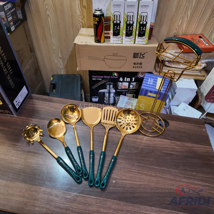 A set of six gold-colored kitchen utensils with dark green handles, including a pasta server, two different ladles, a solid spatula, a slotted spatula, and a slotted spoon.