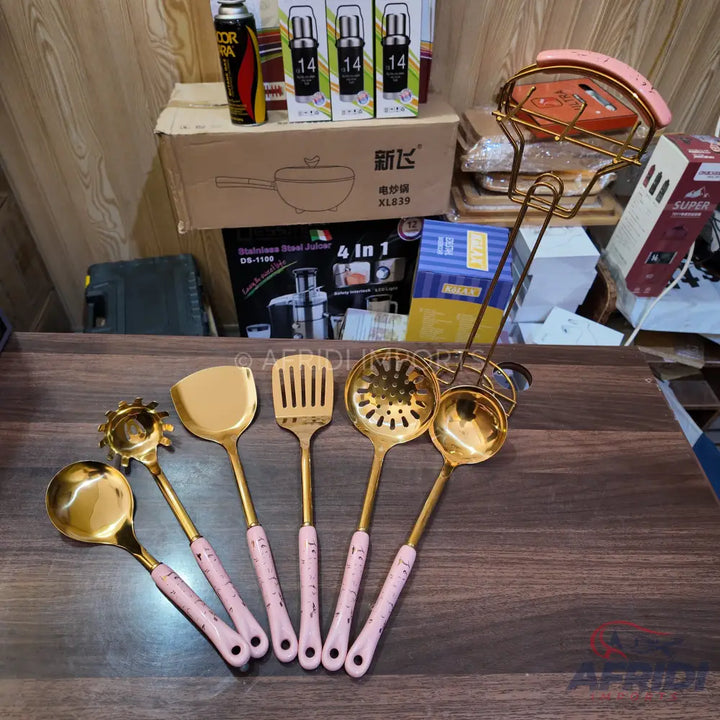 A set of six gold-colored kitchen utensils with light pink handles.