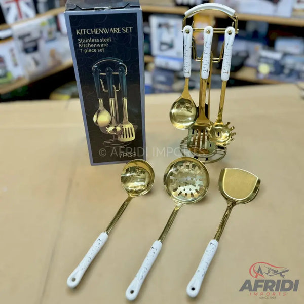 A gold-colored stainless steel kitchenware set with white ceramic handles, including a ladle, slotted spoon, and spatula, with a matching stand.