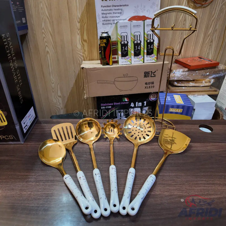 A set of six gold-colored kitchen utensils with white marbled handles, including a ladle, spatula, skimmer, and pasta server, along with a matching golden stand.