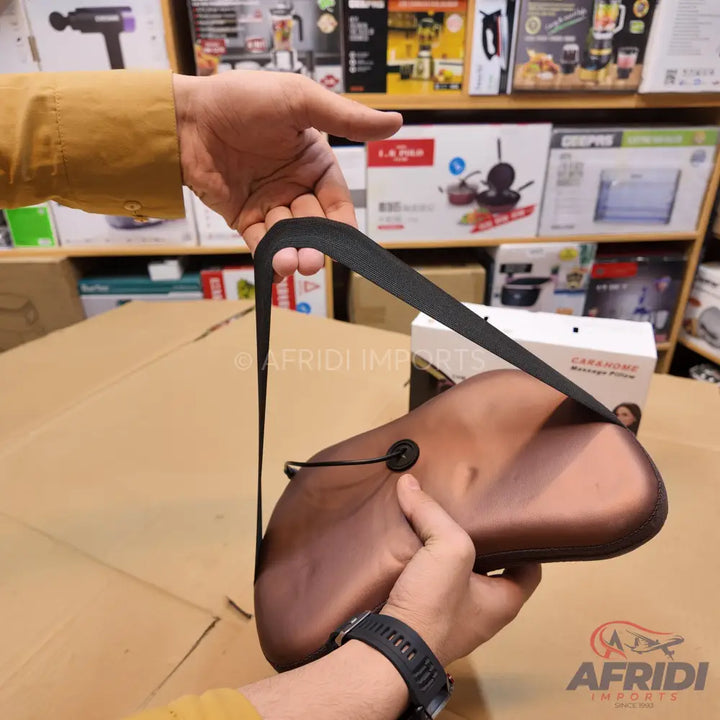 A person holding a brown, ergonomically shaped massage pillow with a black strap and control button.