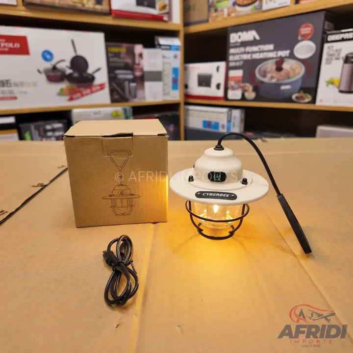 White and black vintage-style portable lantern with a glowing bulb, accompanied by its brown cardboard packaging box and a charging cable.