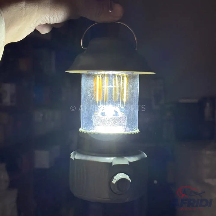 A hand holding a lit, portable, modern-style camping lantern with a dimmer knob.