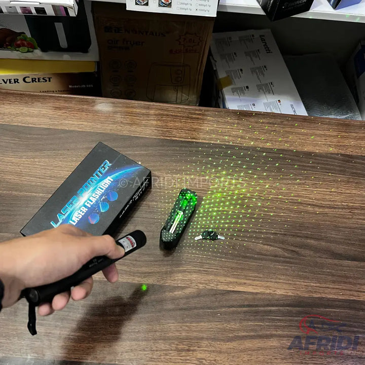 A hand holding a black laser pointer with a red button, next to its box and another green laser pointer.
