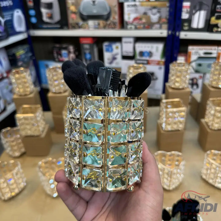 A cylindrical makeup brush holder adorned with clear and light green faceted crystals, set in a gold-toned frame.