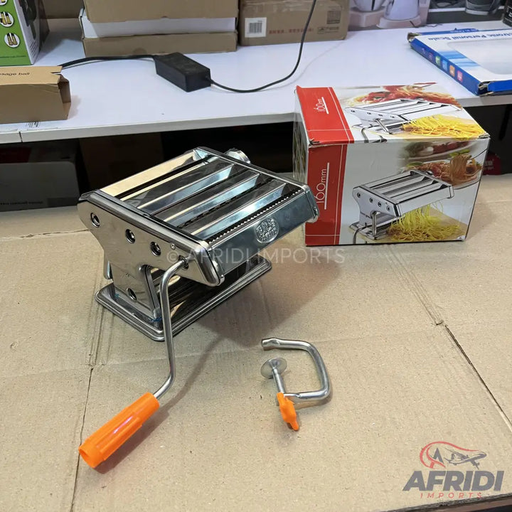 A stainless steel manual pasta maker with an orange handle and table clamp.