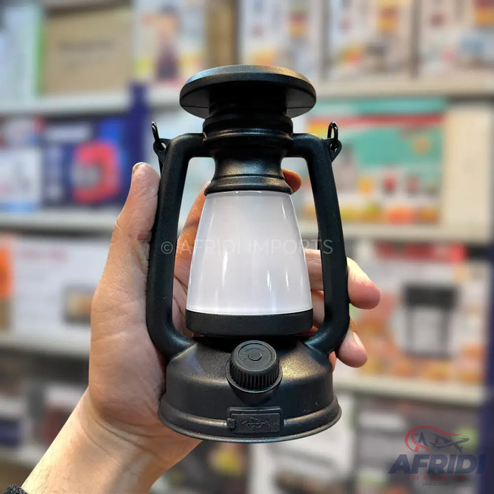 A person’s hand holds a black, modern, battery-powered LED lantern with a white frosted diffuser and a control knob on its base.