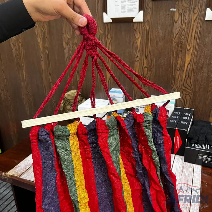 A colorful striped fabric hammock with red support ropes and a wooden spreader bar.