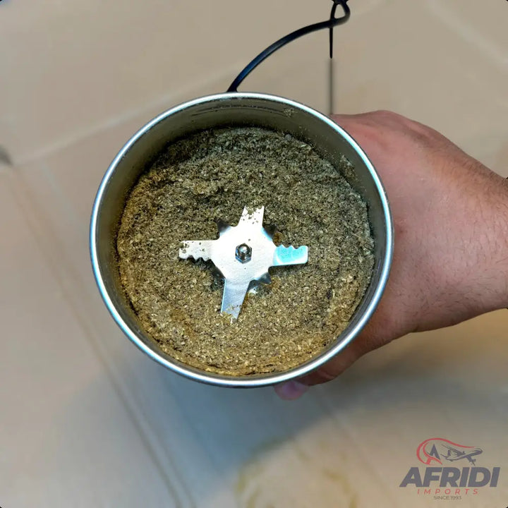 A stainless steel spice grinder with a four-pronged blade, filled with ground green spices.