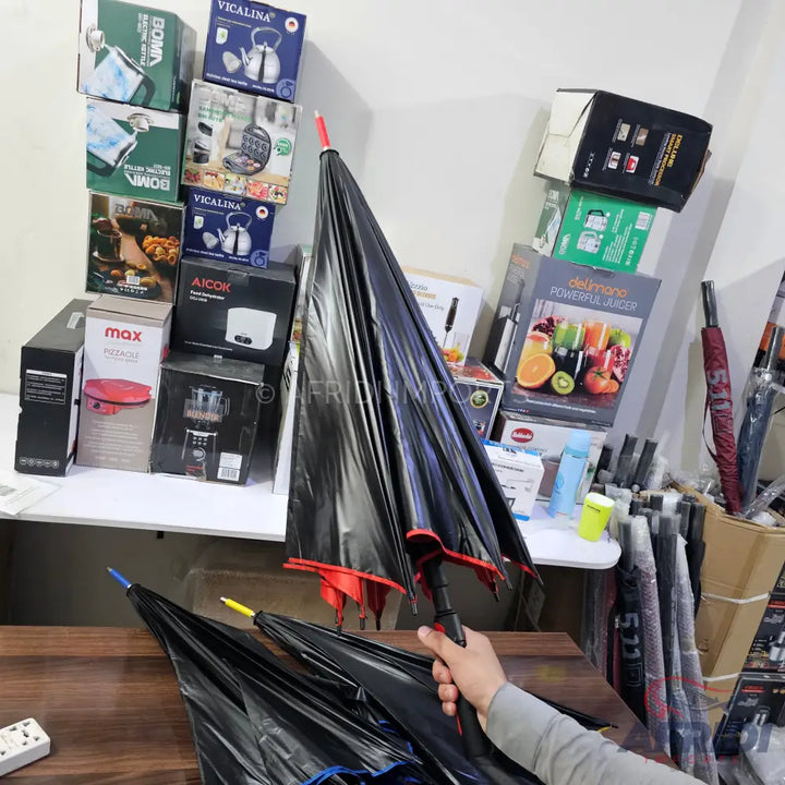 Black umbrella with red accents, held in a hand, surrounded by other umbrellas and boxed household appliances.