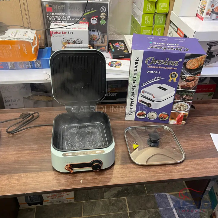 An open, light blue and black electric cooking appliance with a ridged grill on top and a removable glass lid, designed for steaming and frying food.