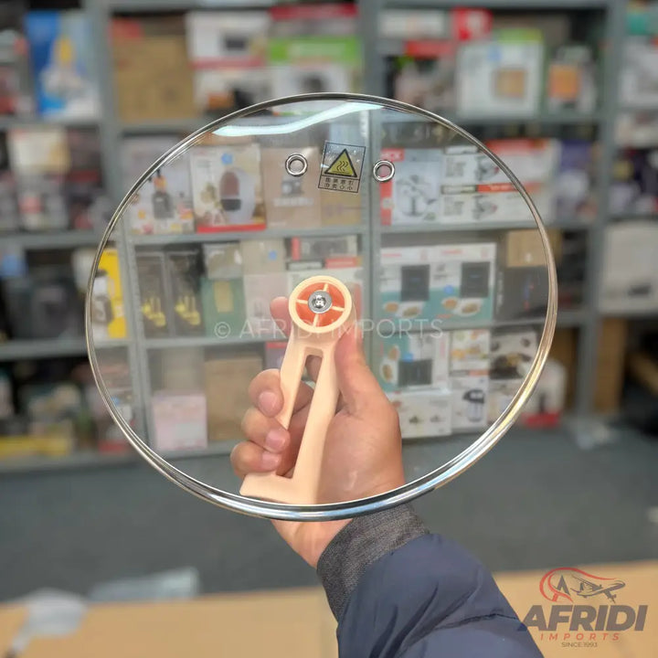 A transparent glass pot lid with a beige and orange foldable handle.