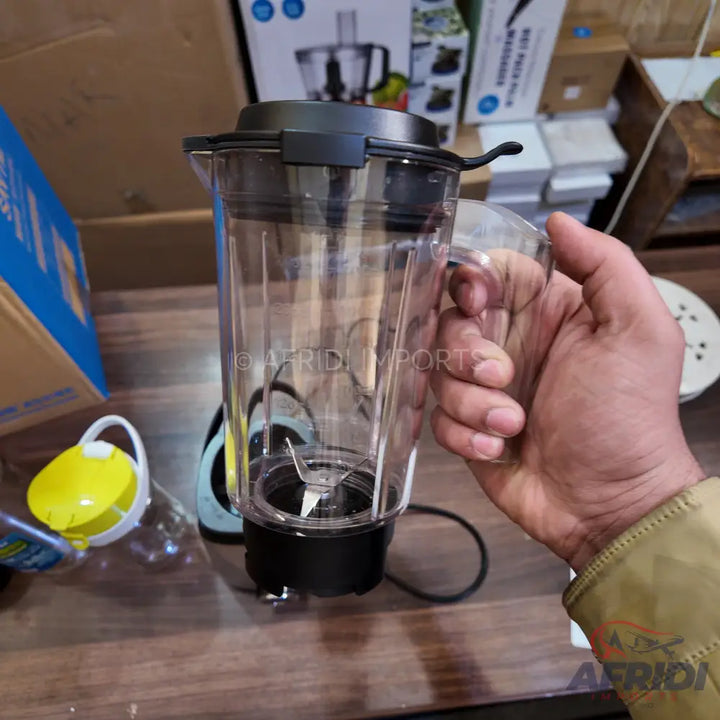 Clear plastic blender pitcher with a black lid and blade assembly, held by a hand.
