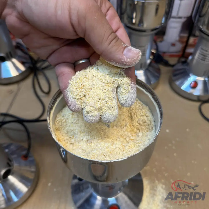 A person’s hand holding a handful of light yellow, coarse-ground flour over a metal grinder filled with more of the same flour.