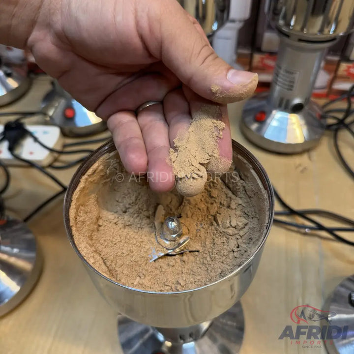 A hand holding a pinch of light brown powder over a stainless steel electric grinder filled with the same powder.