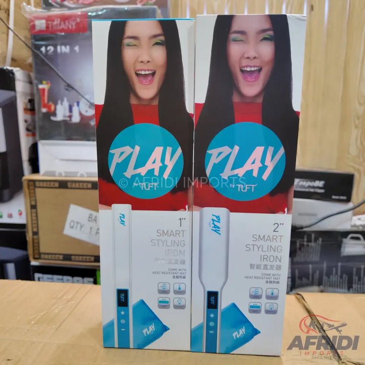 Two boxed ’Play’ brand smart styling irons, one 1-inch and one 2-inch, featuring a woman with long dark hair winking on the packaging.