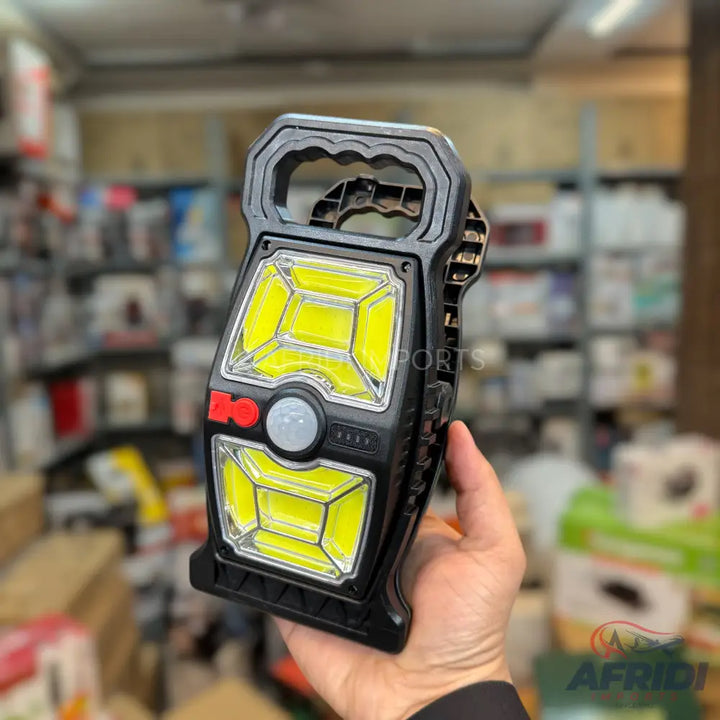 A handheld black work light with a handle, featuring two large rectangular LED panels and a central red button.