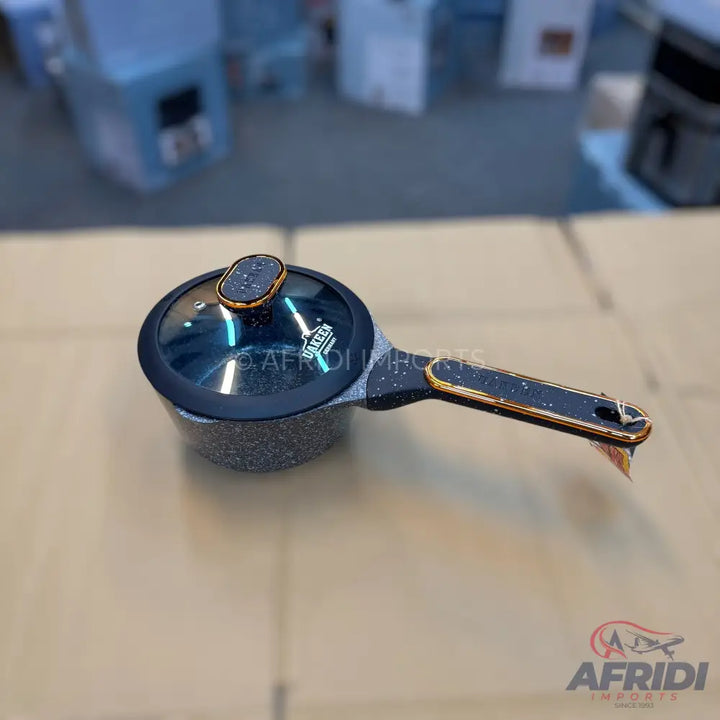 A speckled, dark-colored saucepan with a glass lid and a black handle accented with gold trim.