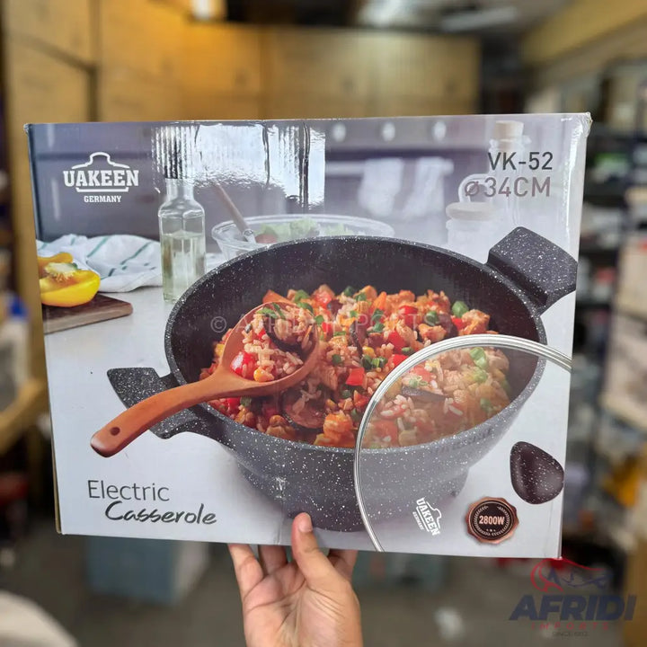 An electric casserole dish with a speckled dark gray finish and a wooden spoon, featuring a glass lid.