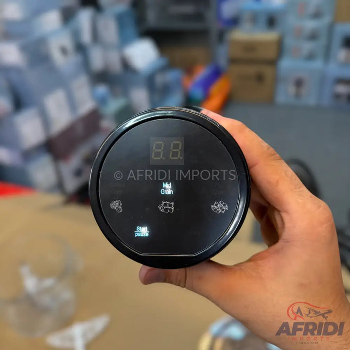A hand holding a black, cylindrical kitchen appliance with a digital display and touch controls for different grind settings.