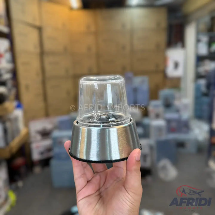 A hand holding a clear glass and stainless steel mini food chopper attachment with blades.