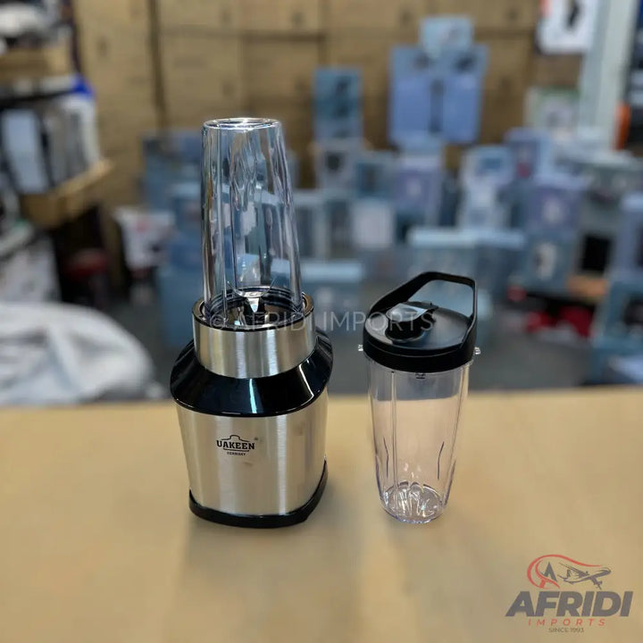 Stainless steel personal blender with two clear blending cups, one with blades and the other with a travel lid.